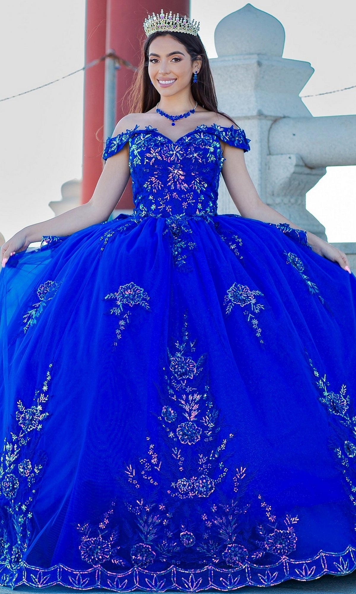 Young woman outdoors in the Quinceanera Ball Gown 8100J with off-the-shoulder design, iridescent beaded details, floral embroidery, lace-up back, and royal blue skirt, smiling as she holds out her dress.